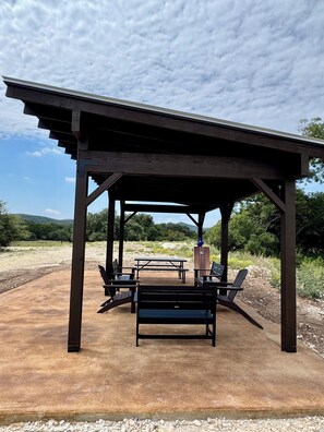 A covered outdoor seating area with benches and a picnic table, surrounded by natural landscape.