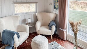 Une pièce chaleureuse avec deux fauteuils, une petite table avec une lampe, une fenêtre donnant sur un beau paysage et un vase contenant des plantes séchées.