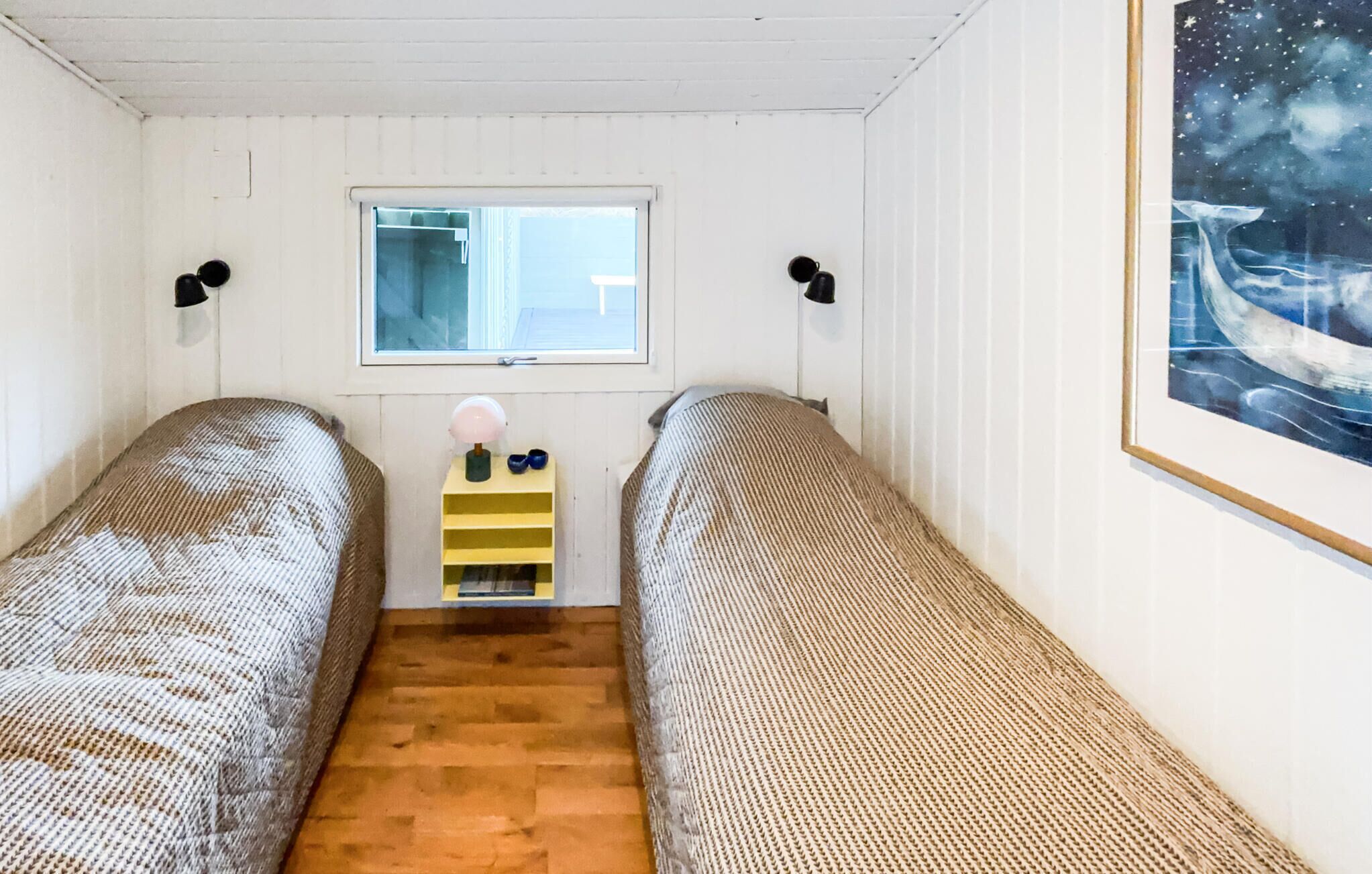 Ein Zimmer mit zwei Einzelbetten, einem Fenster, einem kleinen Tisch mit Lampe und einem gerahmten Bild an der Wand.