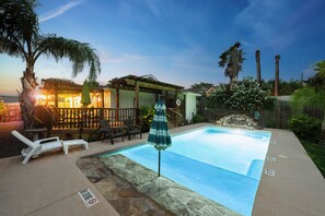 A swimming pool with lounge chairs and an umbrella, surrounded by stone walls and palm trees. - Perfect oasis with private pool and wrap around deck! (Port Aransas)
