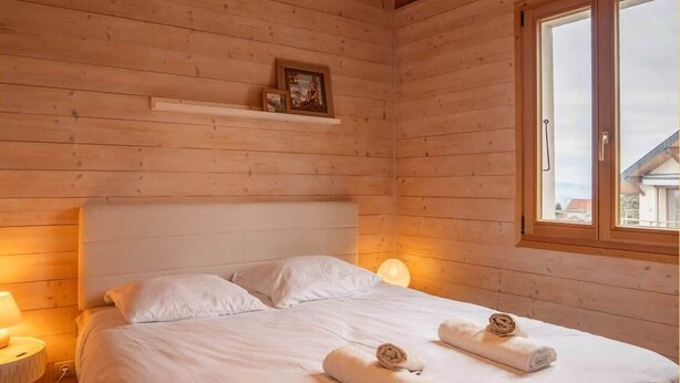 A wooden cabin bedroom with a bed, two pillows, a bedside lamp, and a window. - Maison méserier I T3 I Publier (Publier)
