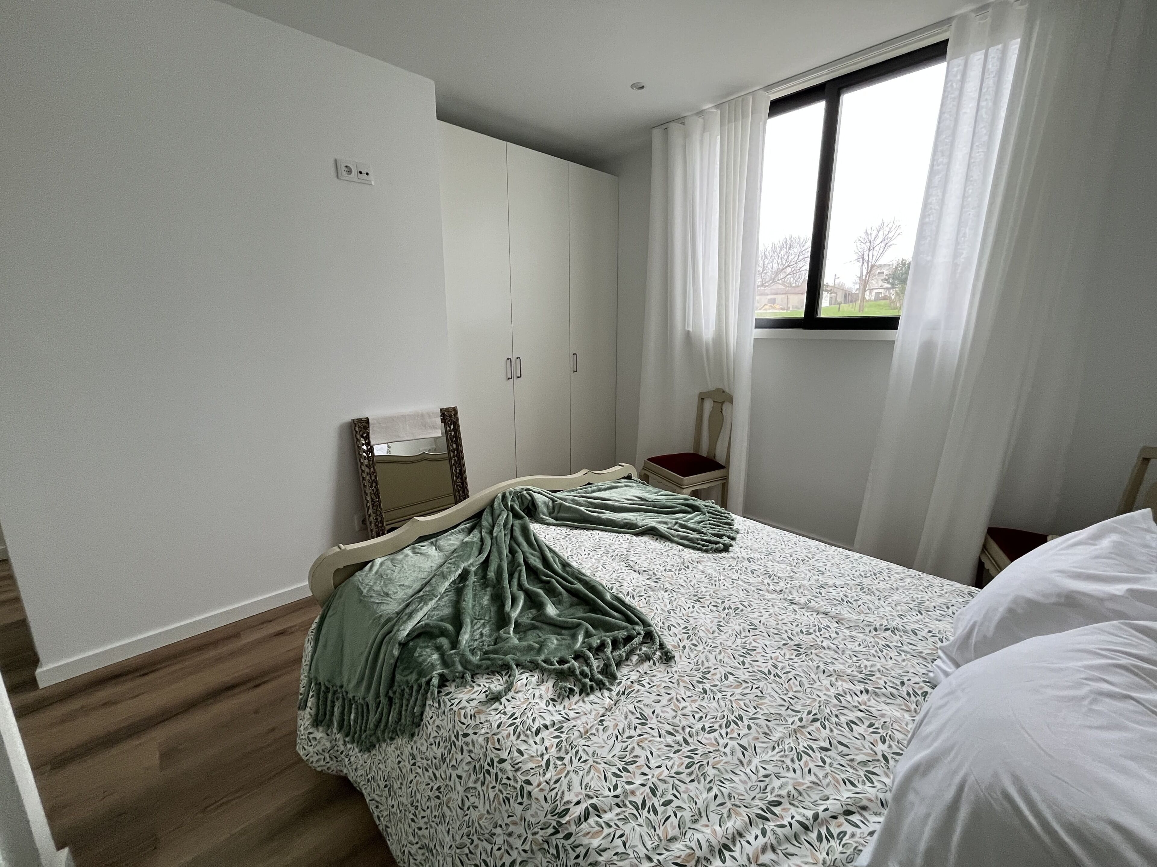 A bedroom with a bed, a chair, a wardrobe, and a window with curtains.