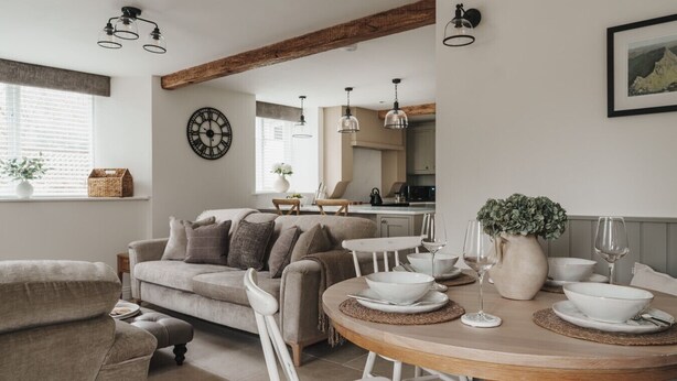 Un salon chaleureux avec un canapé, une table à manger et des chaises, une cuisine en arrière-plan et une horloge au mur.