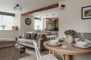 A cozy living room with a sofa, dining table, and chairs, a kitchen in the background, and a clock on the wall. - Valmere Retreat, Luxury Cottage, Windermere (Windermere)