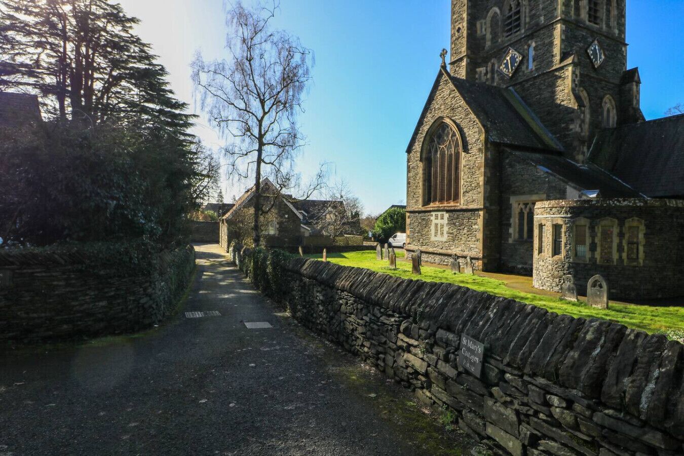 En stensatt gång leder fram till en kyrka med ett högt torn och en mindre byggnad med en skylt där det står ”Graham’s Cottages”.