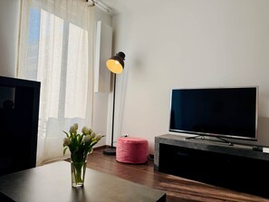 Un salon moderne avec un téléviseur à écran plat, un petit pouf rose, un vase rempli de fleurs et une lampe sur pied.