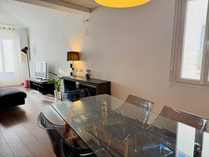 A modern living room with a glass dining table, black chairs, a dark sofa, a TV, and a lamp.