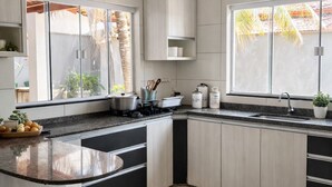 A modern kitchen with a curved island, white cabinetry, and a large window with a view of palm trees. - Vacation Home - Recanto Flor de Laranjeira, Palmas-Tocantins. (Palmas)