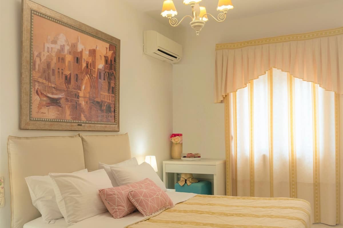 Un dormitorio con cama, mesita de noche, lámpara, un cuadro y ventana con cortinas.
