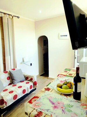 Un soggiorno con divano, televisore, tavolo da pranzo con tovaglia fiorata, una bottiglia di vino e una ciotola di frutta.