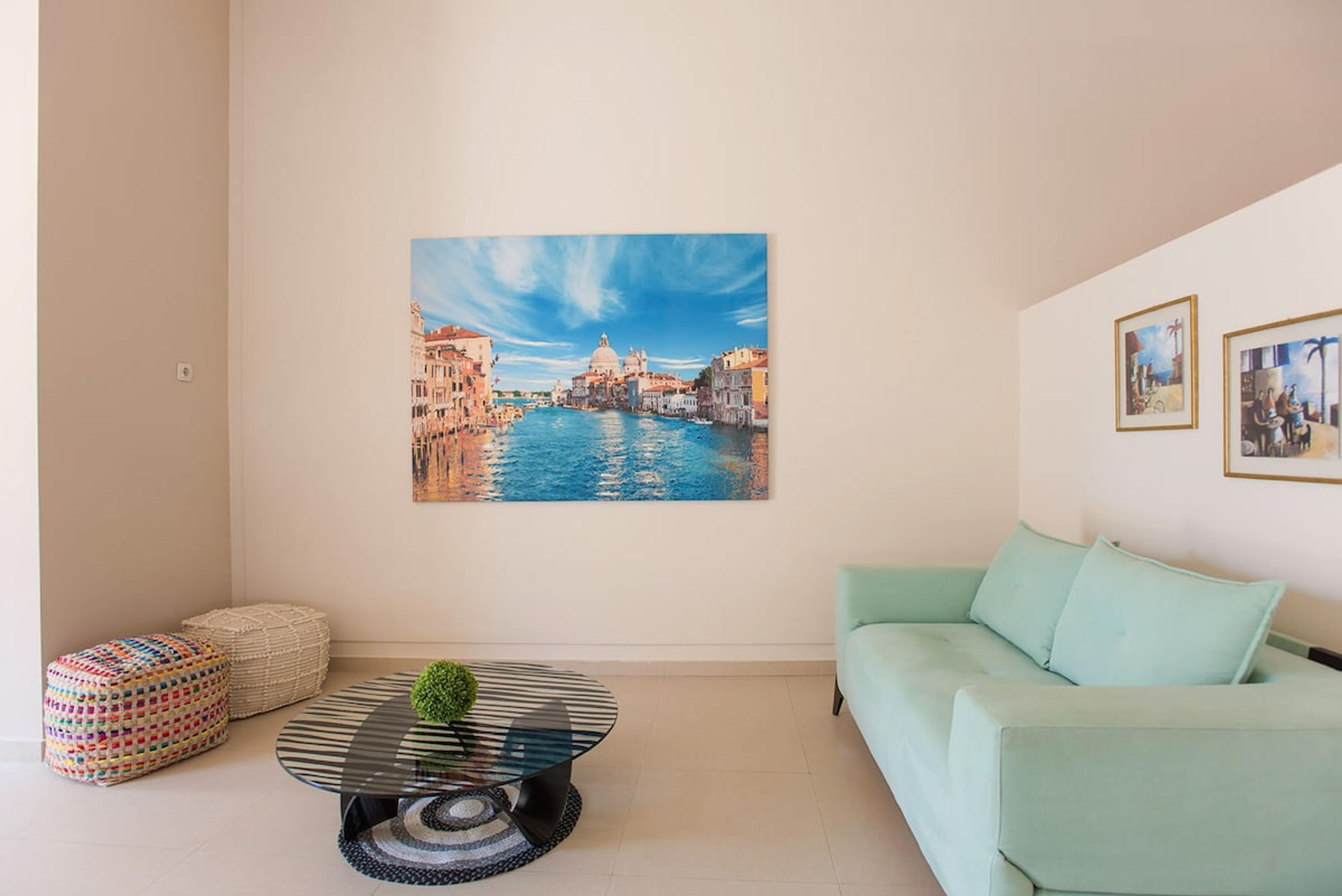 Een woonkamer met een groot schilderij van een gracht, een ronde glazen salontafel, een lichtgroene bank en twee poefs.