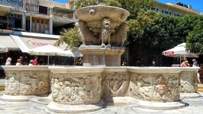 Ein Steinkunstbrunnen mit feinen Schnitzereien, umgeben von einem Platz mit Geschäften und Außenbestuhlung.