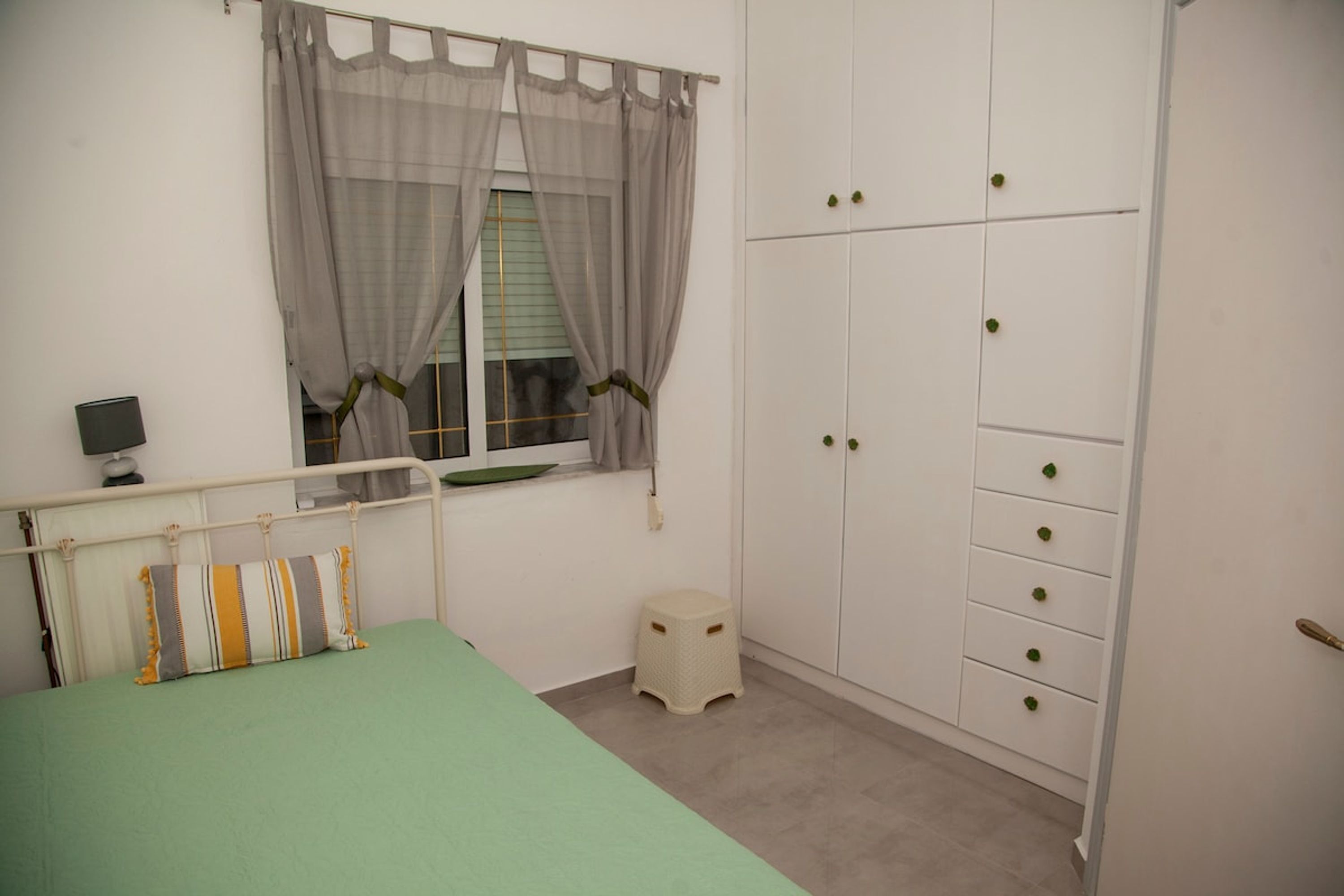 Quarto com cama, janela com cortinas, guarda-roupa com várias gavetas e um banquinho pequeno.