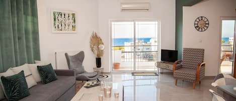 A living room with a sofa, armchair, coffee table, and a view of the sea through a glass door.