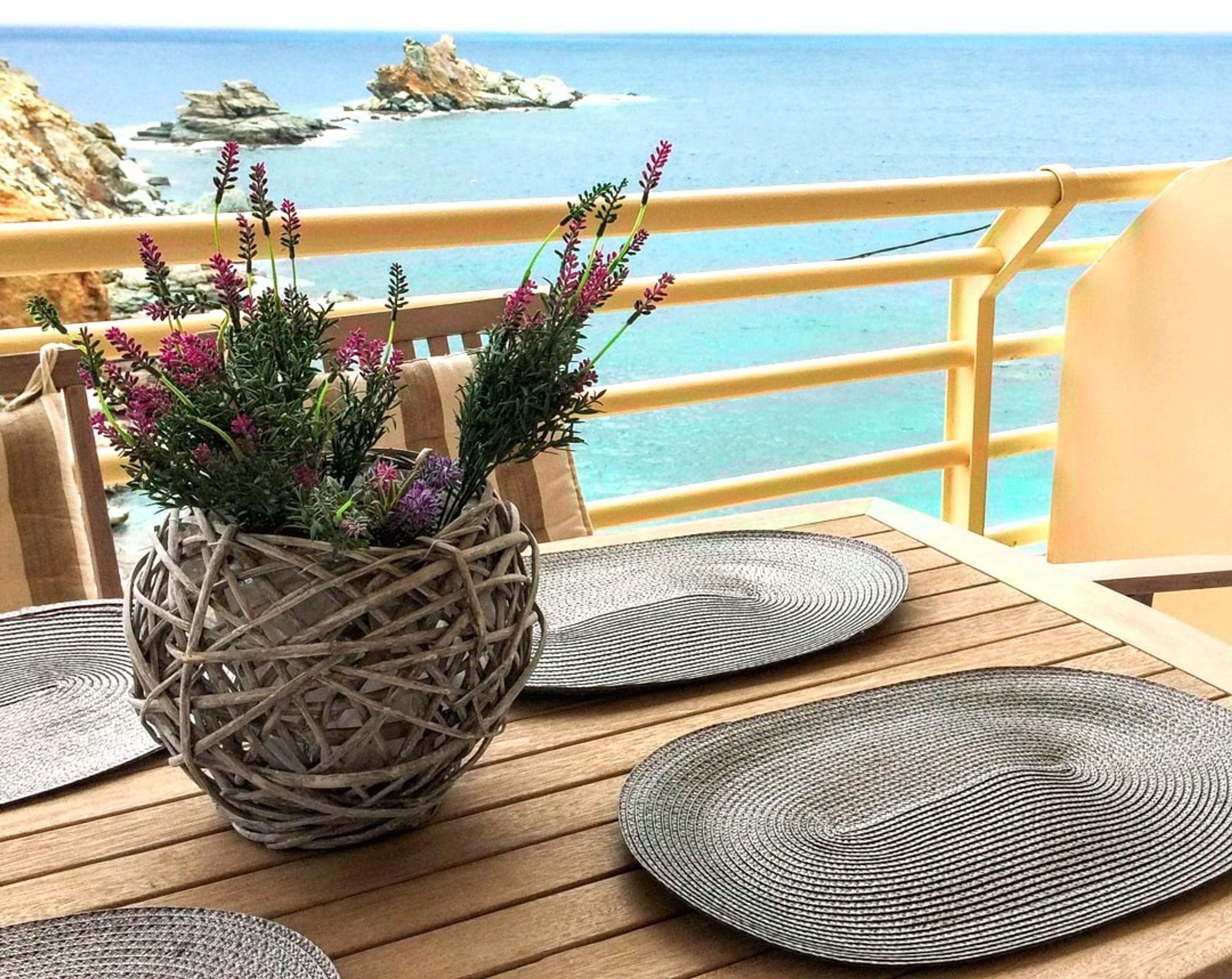Un panier en osier orné de fleurs violettes, posé sur une table en bois recouverte de sets de table en fibres tressées, d’où l’on peut admirer une vue sur la côte.