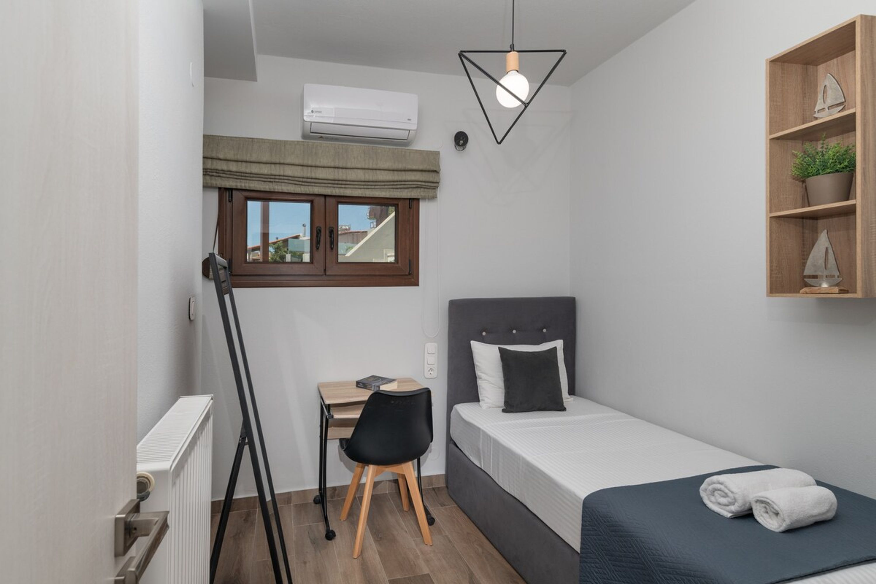 A compact room with a bed, a desk, a chair, a radiator, an air conditioning unit, a window with a curtain, a hanging light fixture, and a shelf with a plant.