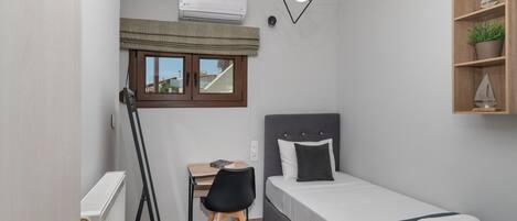 A compact room with a bed, a desk, a chair, a radiator, an air conditioning unit, a window with a curtain, a hanging light fixture, and a shelf with a plant.