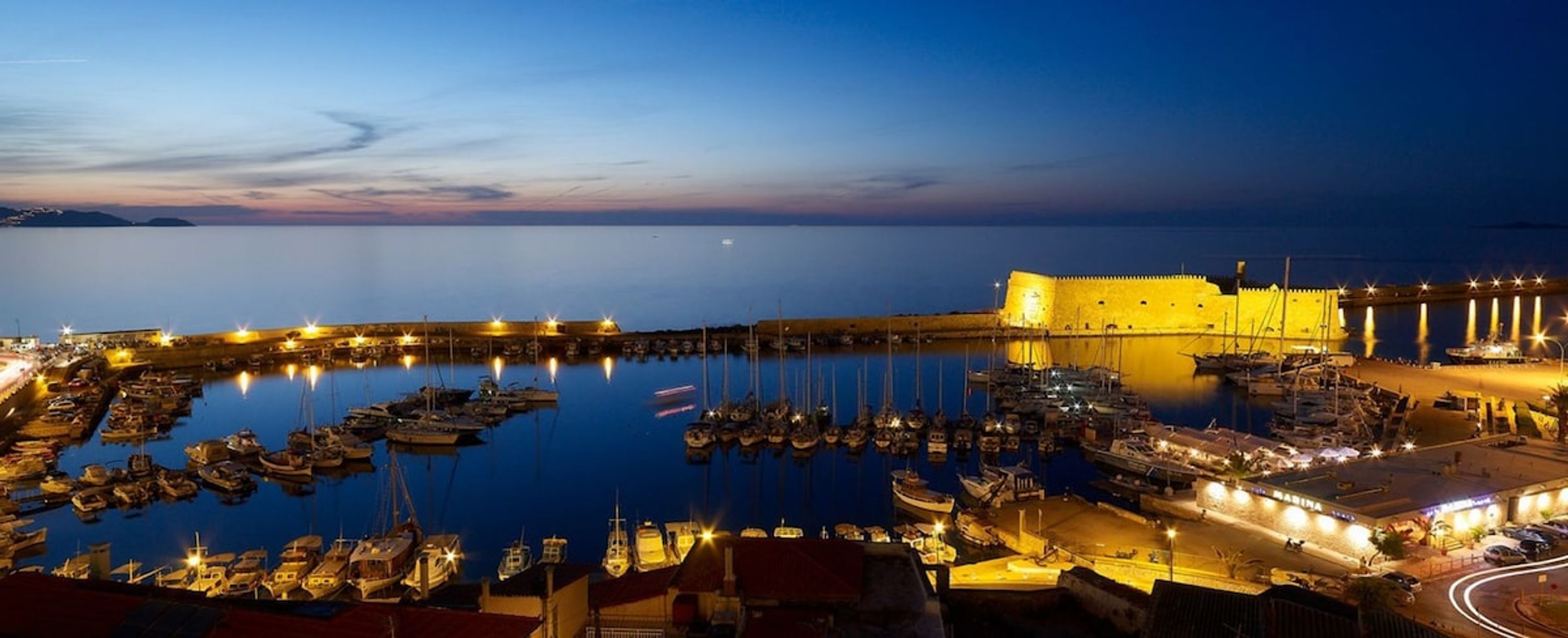 A harbor at dusk with boats docked, a fortified building, and a marina with lights.
