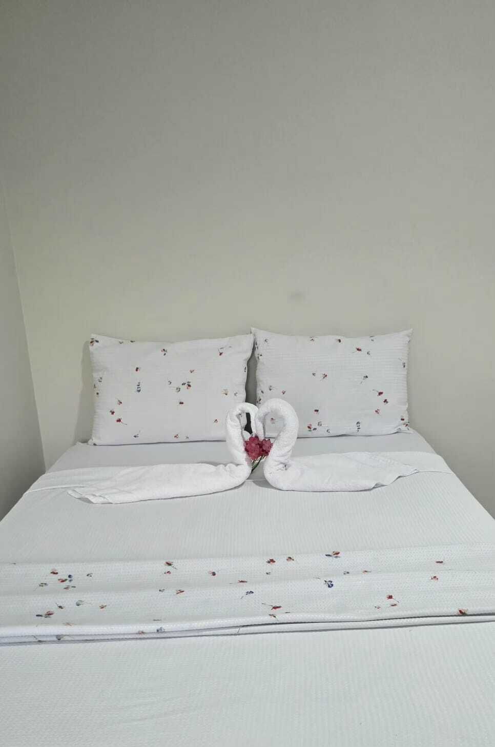 Una cama con ropa de cama blanca y dos almohadas, un arreglo de toallas con forma de corazón y una pequeña flor rosa.