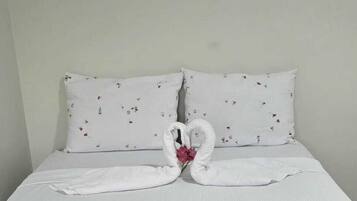 Una cama con ropa de cama blanca y dos almohadas, un arreglo de toallas con forma de corazón y una pequeña flor rosa.