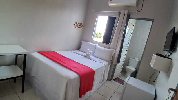 Un lit simple avec une couverture rouge, une petite table, un miroir et un téléviseur.