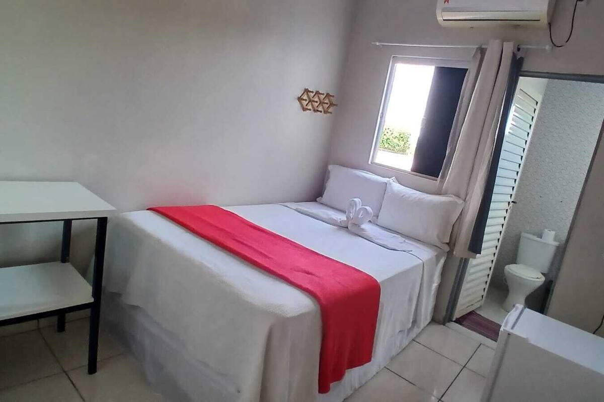 A single bed with a red blanket, a small table, a mirror, and a TV.