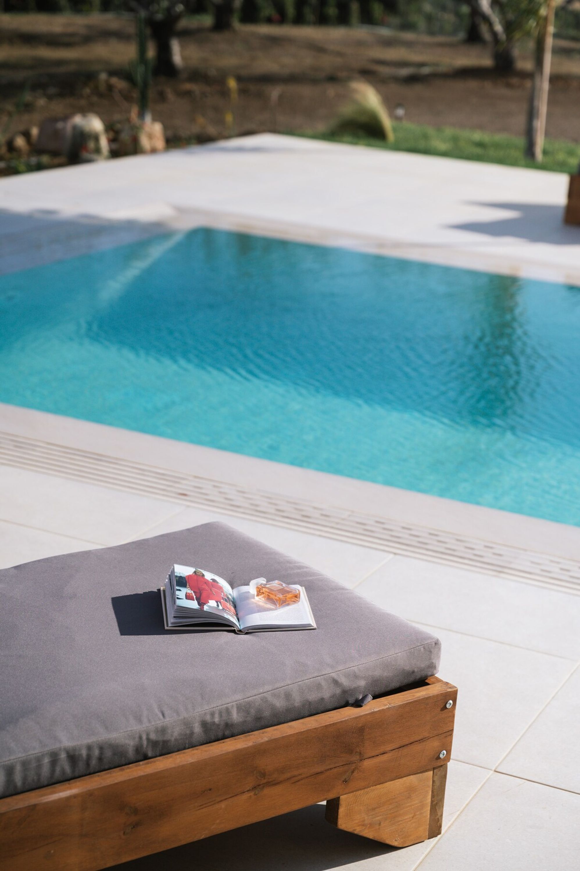A wooden bench with a cushion and a magazine, overlooking a swimming pool.