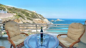 A balcony with wicker chairs and a glass table, overlooking a coastal landscape with rocky terrain and the sea. - Eleni Sea Luxury Apartment (Malevizi)