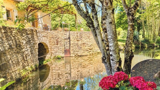 A stone bridge over a calm water body, surrounded by greenery and a building with a tiled roof. - Cozy cottage in the grounds of an old mill (Payrignac)
