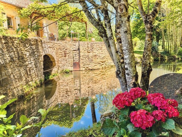 Un pont de pierre enjambant une étendue d’eau paisible, entouré de verdure et bordé d’un bâtiment dont le toit est recouvert de tuiles.