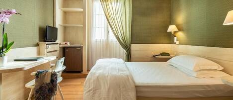 A hotel room with a bed, a desk with a TV, a chair, a lamp, and a window with curtains.