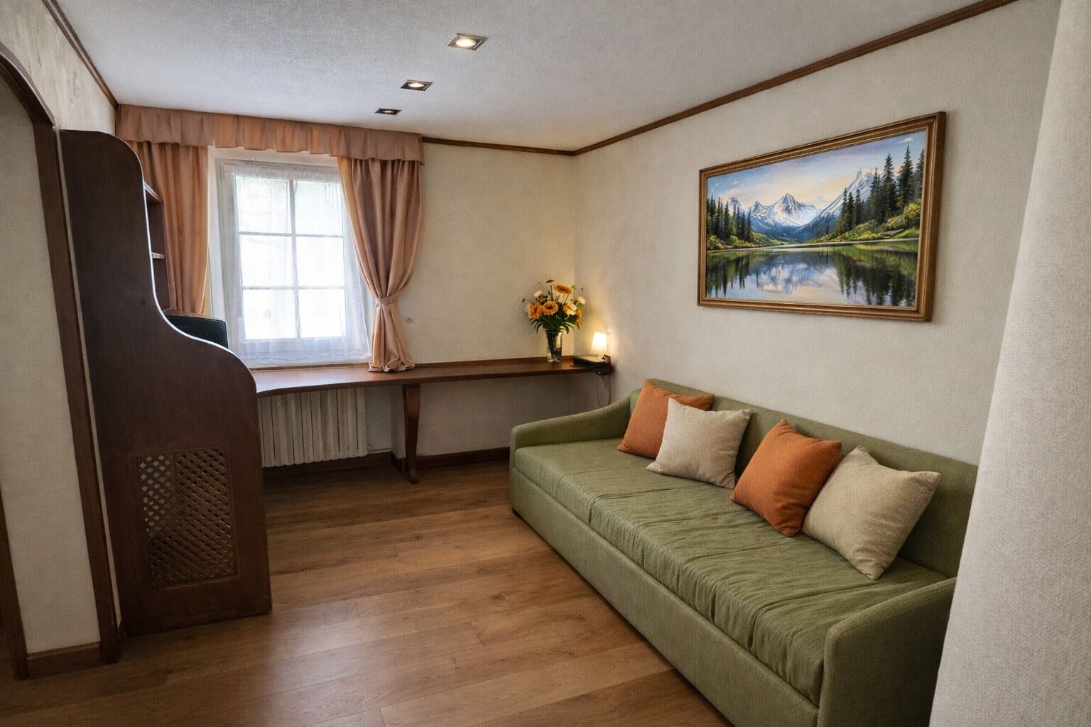 A room with a wooden floor, a green sofa with cushions, a window with curtains, a painting on the wall, and a wooden bench.