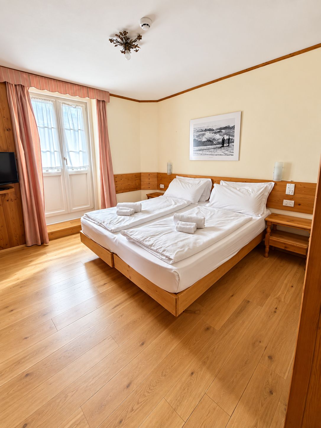 A hotel room with a wooden bed, a television, a painting on the wall, and a window with curtains.