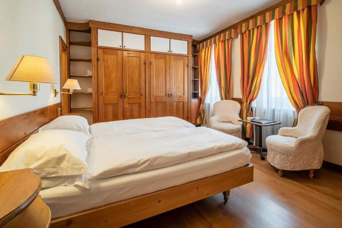 Ett hotellrum med en säng i trä, två stolar, ett litet bord och en garderob med flera dörrar.