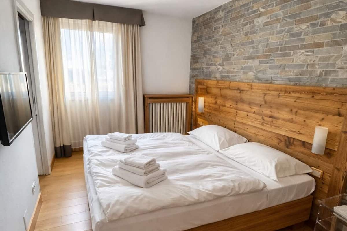 A hotel room with a wooden bed, a stone wall, a ceiling light fixture, and a window with curtains.