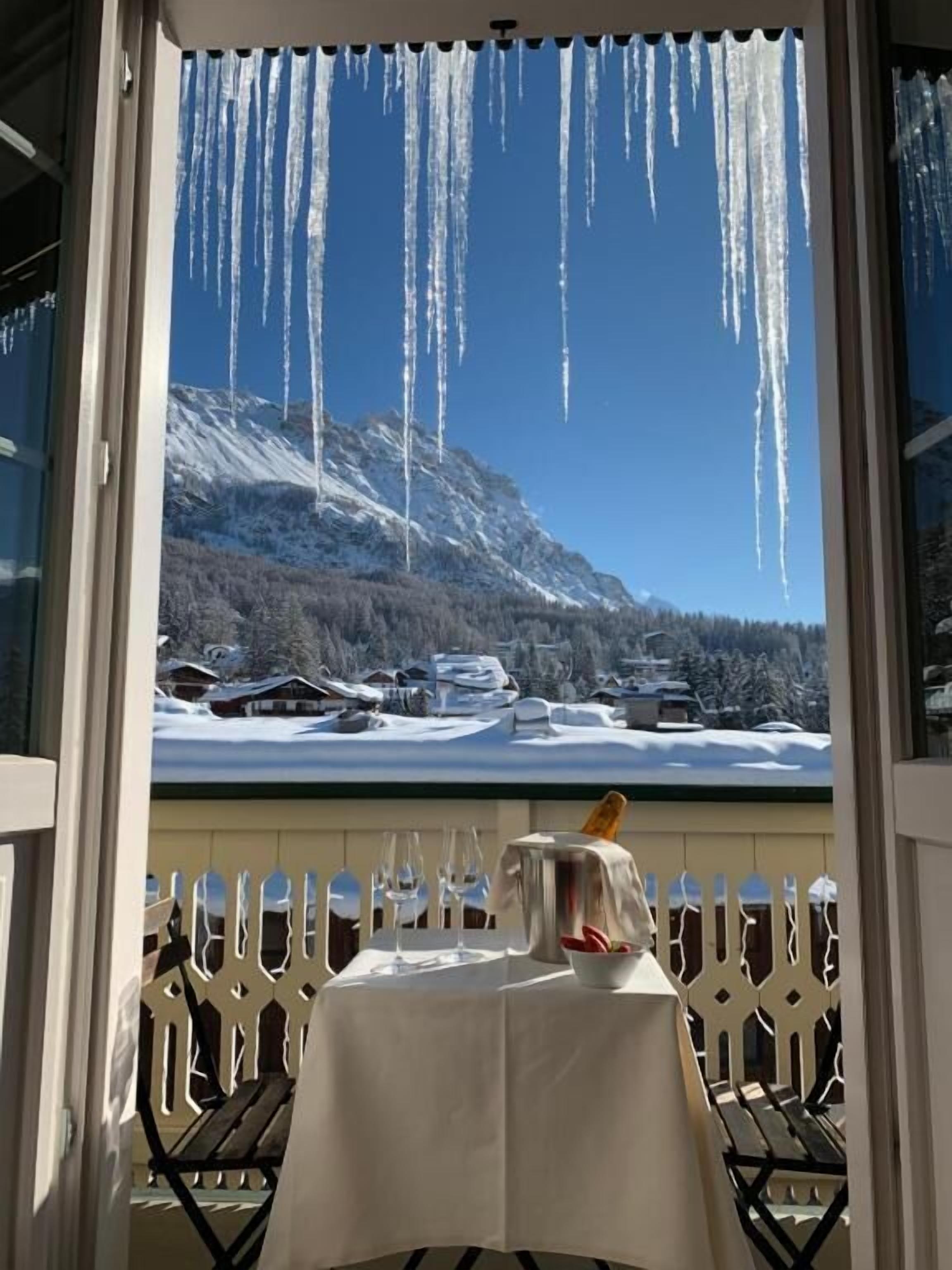 A table with a bottle and glasses, a view of a snowy mountain landscape through a window, and icicles hanging from the window frame.