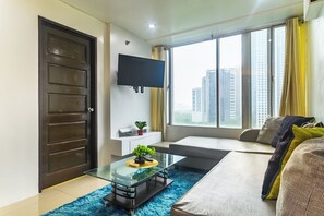 A modern living room with a glass coffee table, a sofa, a TV mounted on the wall, and a large window with a view of buildings. - Cozy City Stays at Seibu Tower (Taguig)