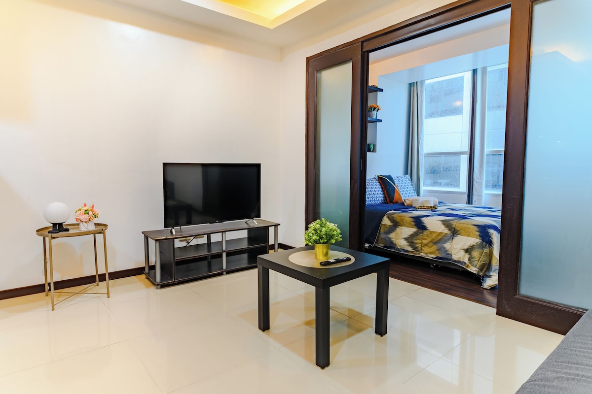 A modern living room with a flat-screen TV, a small table with a plant, and a bed with a patterned bedspread.