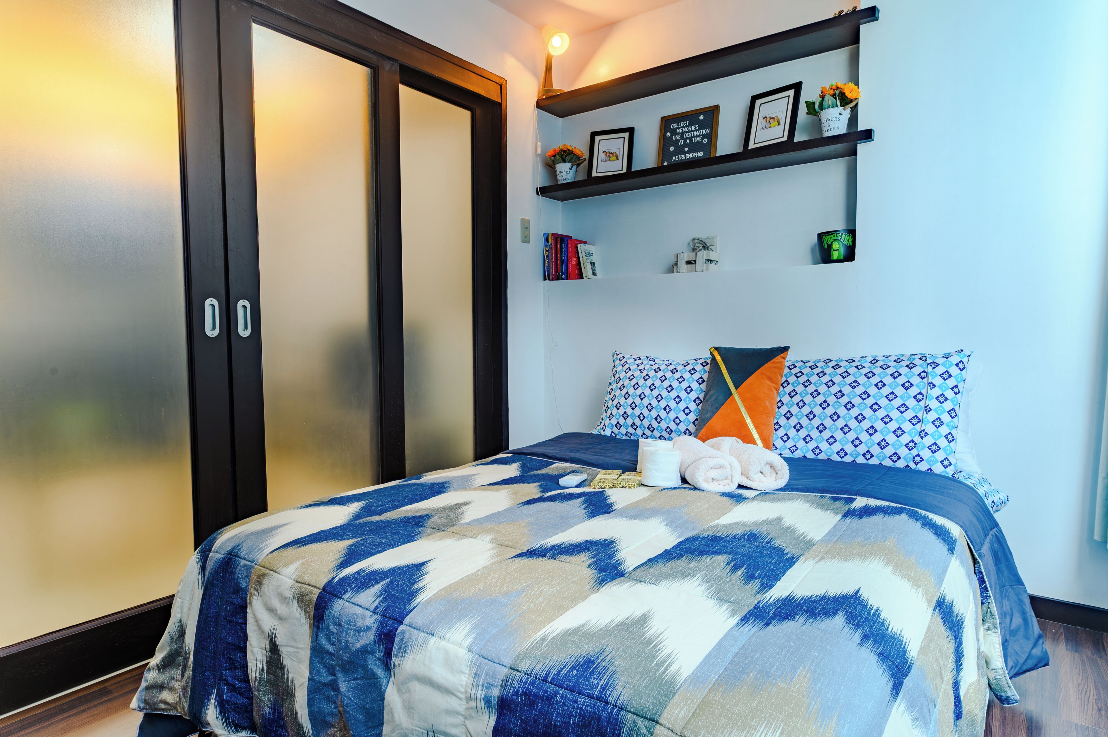 A hotel room with a bed, a wardrobe, a shelf with decorative items, and a wall with a framed picture.