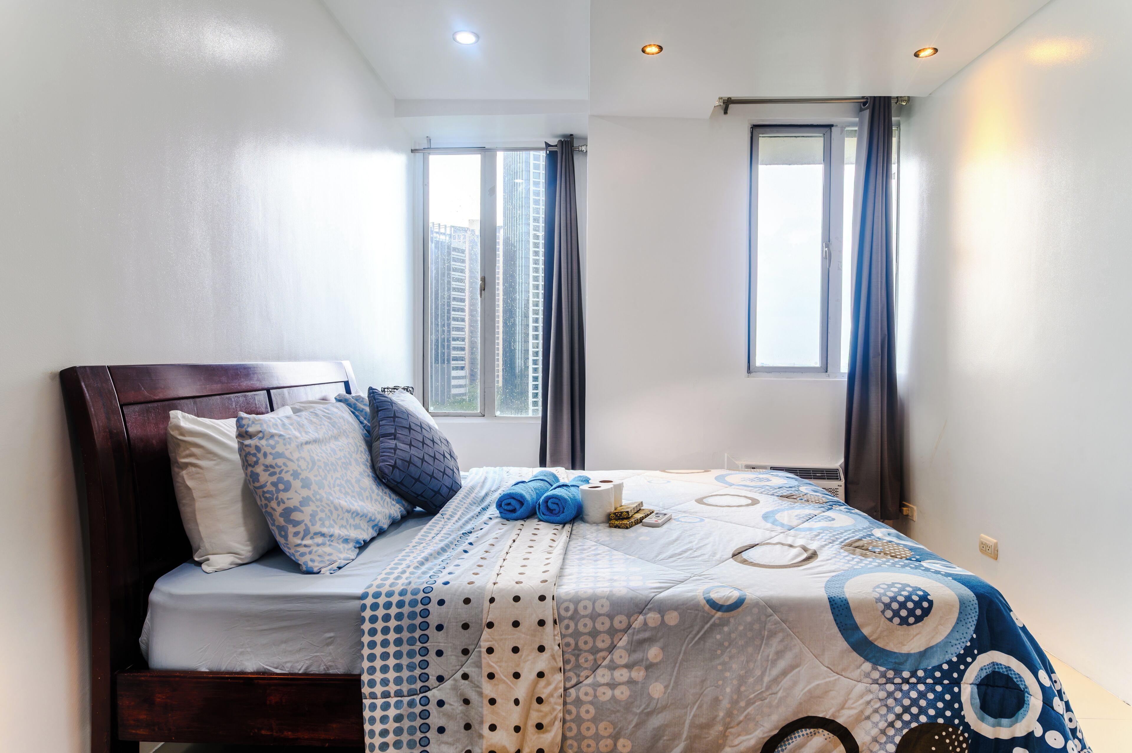 A neatly made bed with a patterned bedspread, pillows, and a rolled towel. There are two windows with curtains, a ceiling light, and a wall-mounted air conditioning unit.