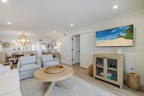 Ein modernes Wohnzimmer mit einem runden Couchtisch, hellblauen Sofas, einem Fernseher, der eine Strandansicht zeigt, und einer Holzkommode mit Glastüren.