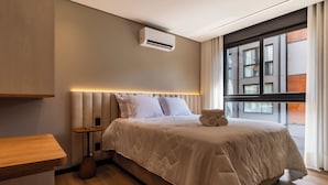 Une chambre d’hôtel moderne dotée d’un grand lit, d’une table de chevet, d’une petite table, d’un appareil de climatisation et d’une fenêtre donnant sur des immeubles.