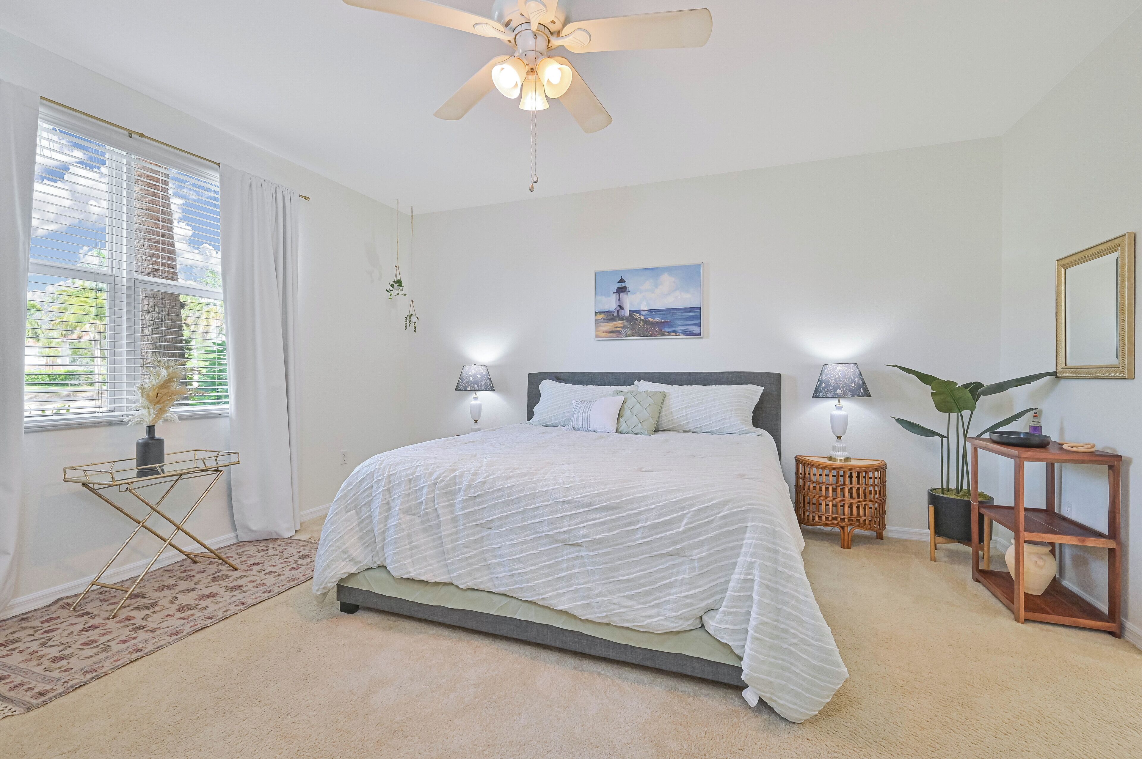 A bedroom with a bed, ceiling fan, bedside lamps, a painting, a plant, and a window with curtains.
