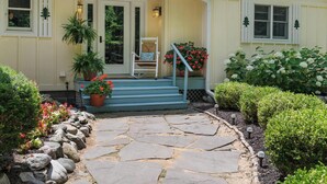 Une maison avec un chemin de pierre menant à un porche doté de marches, d’une chaise et de plantes en pot.