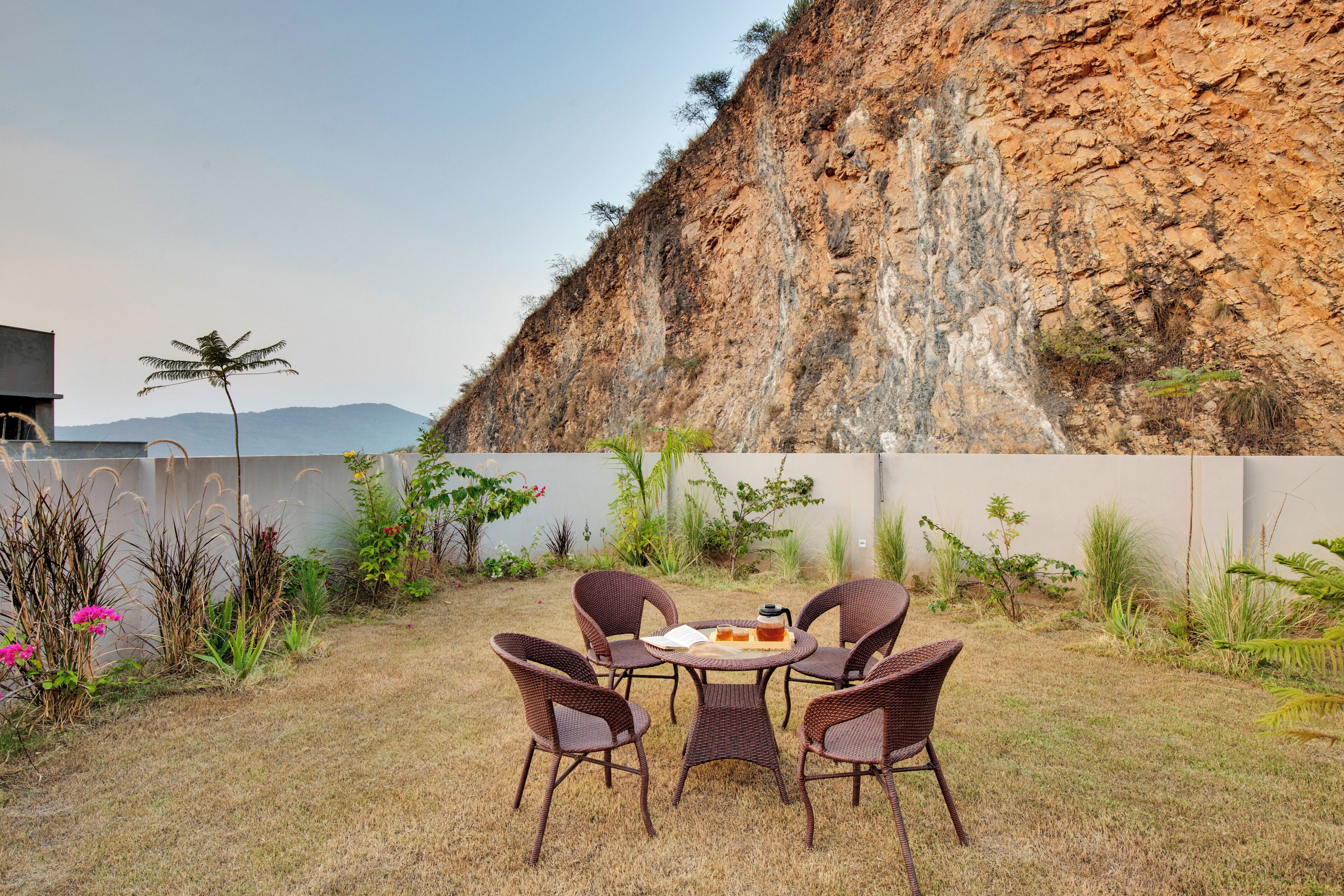 A patio with wicker chairs and a table, surrounded by plants and a view of a rocky cliff.