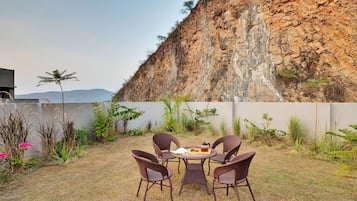 A patio with wicker chairs and a table, surrounded by plants and a view of a rocky cliff.