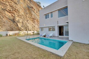 Una casa moderna con piscina, dos sillones de descanso y vistas a una ladera rocosa.