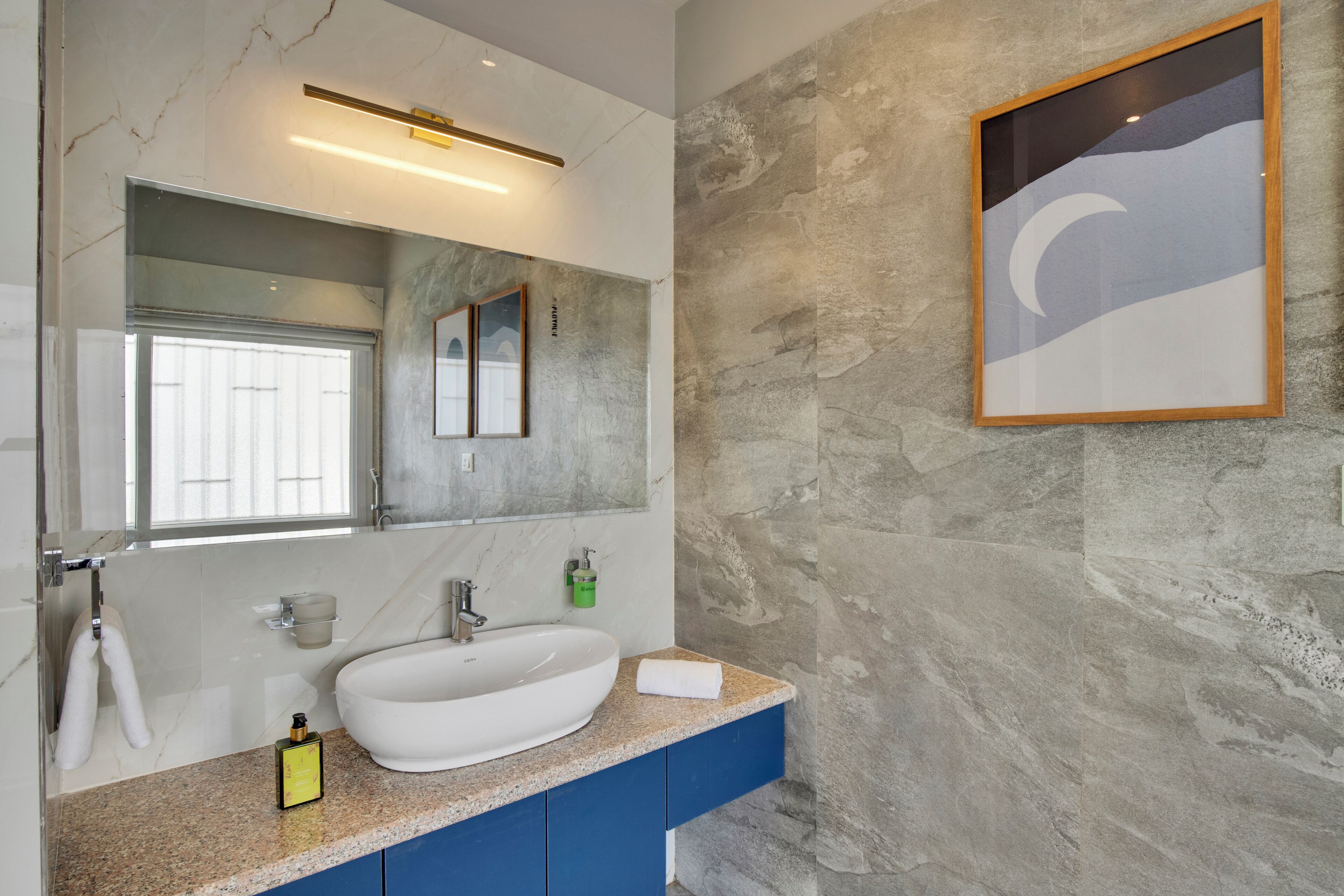 Une salle de bain avec un lavabo blanc, des meubles bleus, un grand miroir et une œuvre d’art encadrée accrochée au mur.