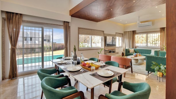 A dining area with a table set for four, green upholstered chairs, and a view of a swimming pool. - ELIVAAS 3 BHK Villa Shourya Bliss (Udaipur)
