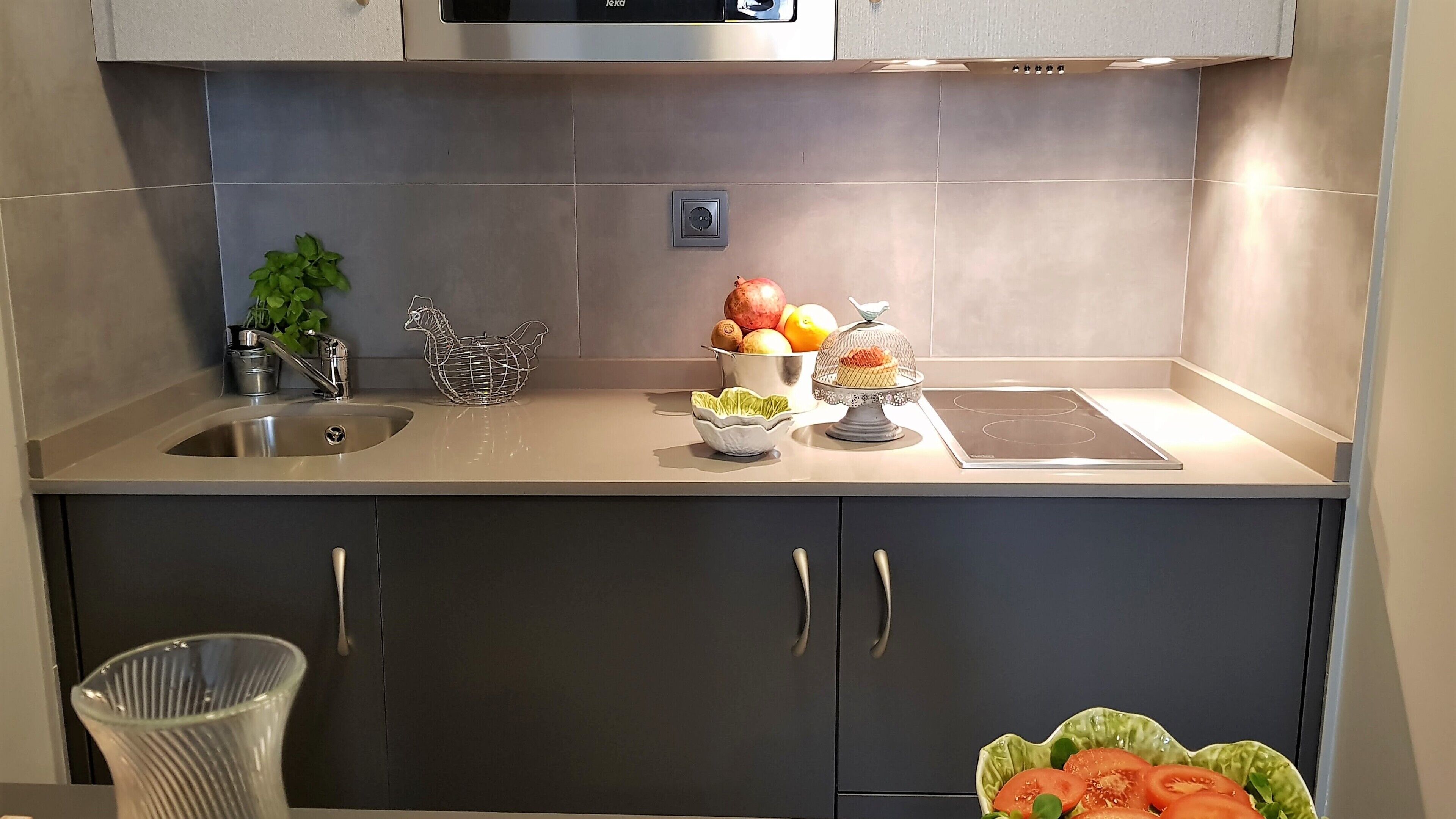 A modern kitchen with a stainless steel sink, a kettle, a fruit bowl, and a bowl of tomatoes.